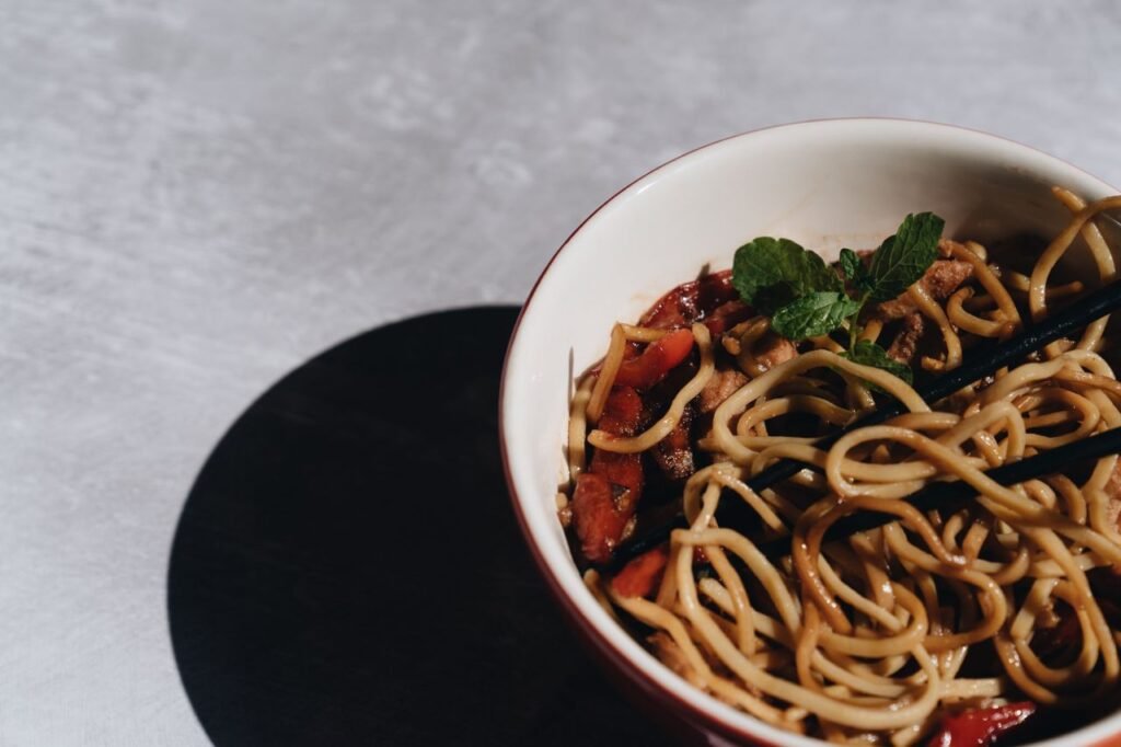 Bowl of Asian noodles with black chopsticks, seasoned vegetables, and fresh mint garnish.